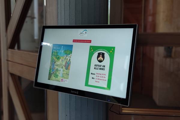 Digitaler Informationsbildschirm in einer Kirche, der eine Einladung zur Veranstaltung 'Aufruf an alle Minis' und ein Plakat mit religiösem Motiv anzeigt.