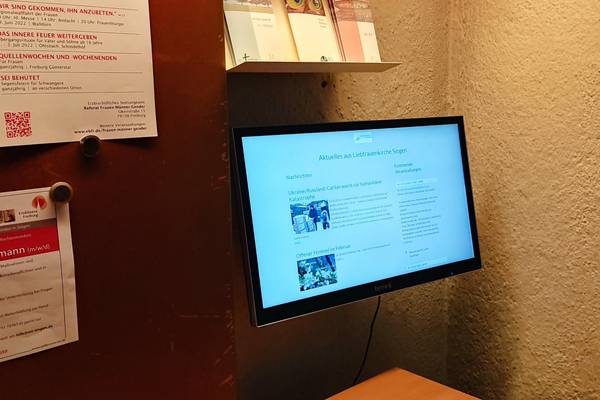 Bildschirm mit aktuellen Informationen der Liebfrauenkirche Singen, umgeben von Aushängen und Broschüren in einem Kirchenflur.