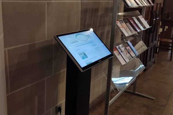 Interaktiver Informationsbildschirm in einer Kirche, umgeben von Prospektständern mit Broschüren, stehend vor einer Steinwand.