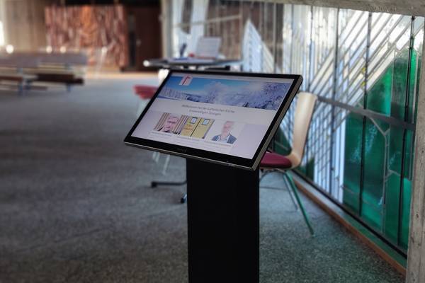 Digitaler Kiosk in einem modernen Kirchenraum mit Glasfenstern, der Besuchern Informationen zur katholischen Kirche auf einem Bildschirm anzeigt
