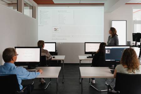 Frau hält Schulung mit Präsentation vor Teilnehmern an Computerarbeitsplätzen