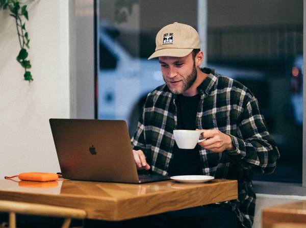 Quelle: kal visuals unsplash Mann sitzt an einem Tisch, arbeitet am Laptop und hält eine Tasse Kaffee in der Hand